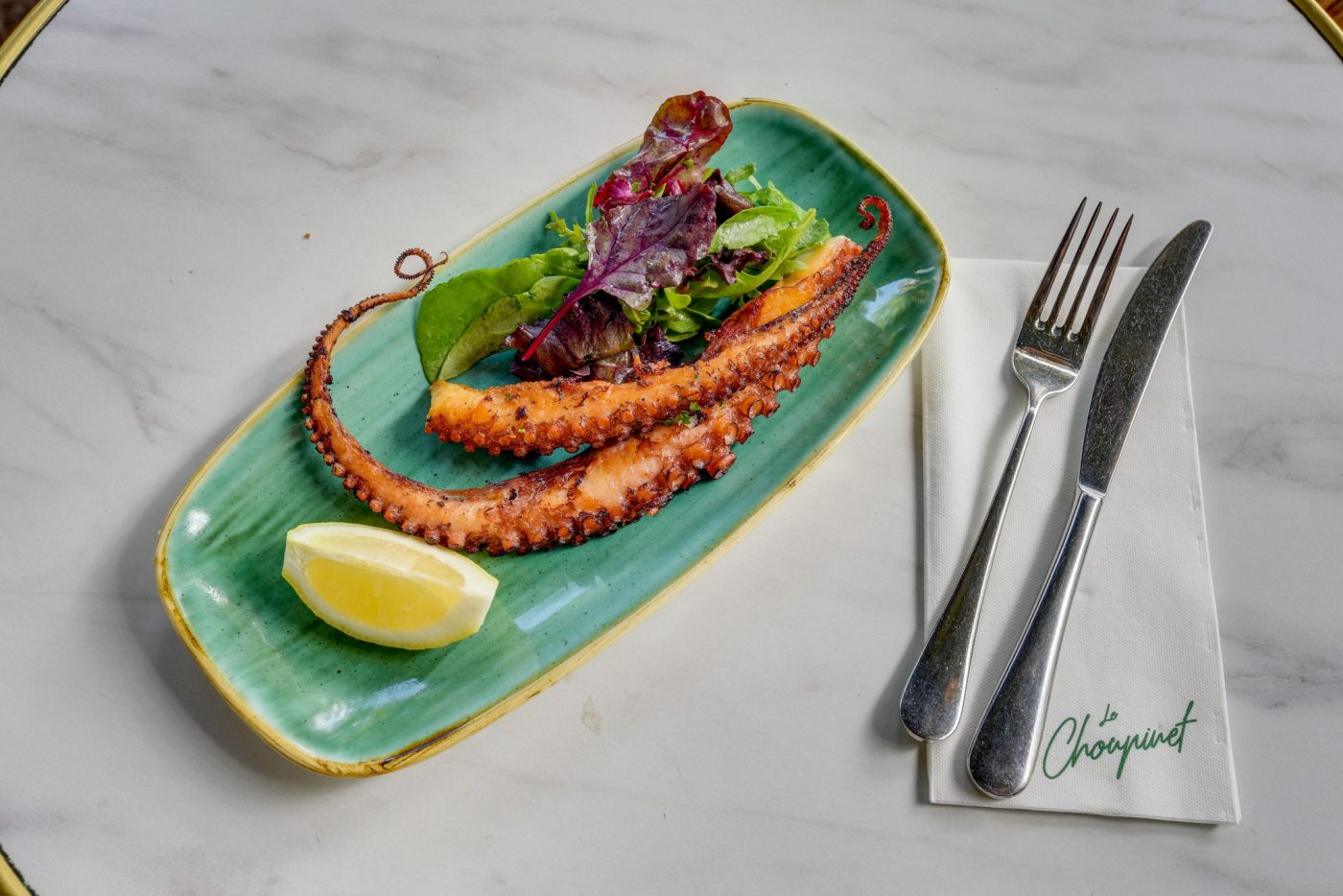 Poulpe grillé dans une assiette bleue/verte déstructurée avec un quart de citron et quelques feuilles de salade verte