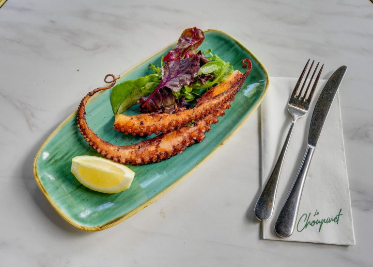 Poulpe grillé dans une assiette bleue/verte déstructurée avec un quart de citron et quelques feuilles de salade verte